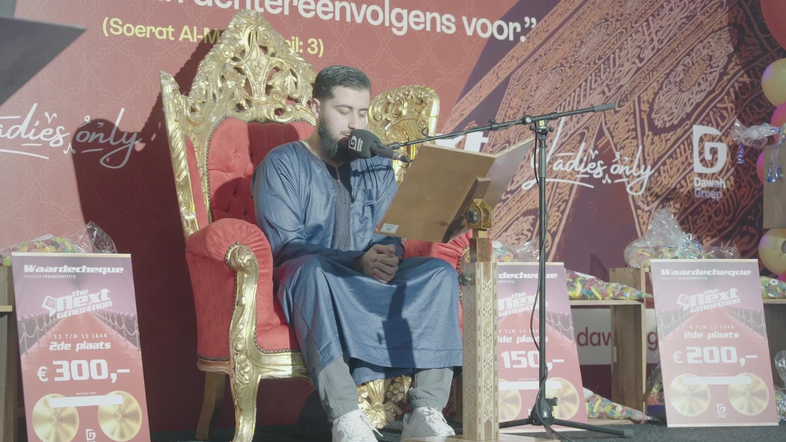 Een man reciteert vanaf een rood met gouden troon op het podium, still uit de aftermovie van de Qurancompetitie van Dawah Groep