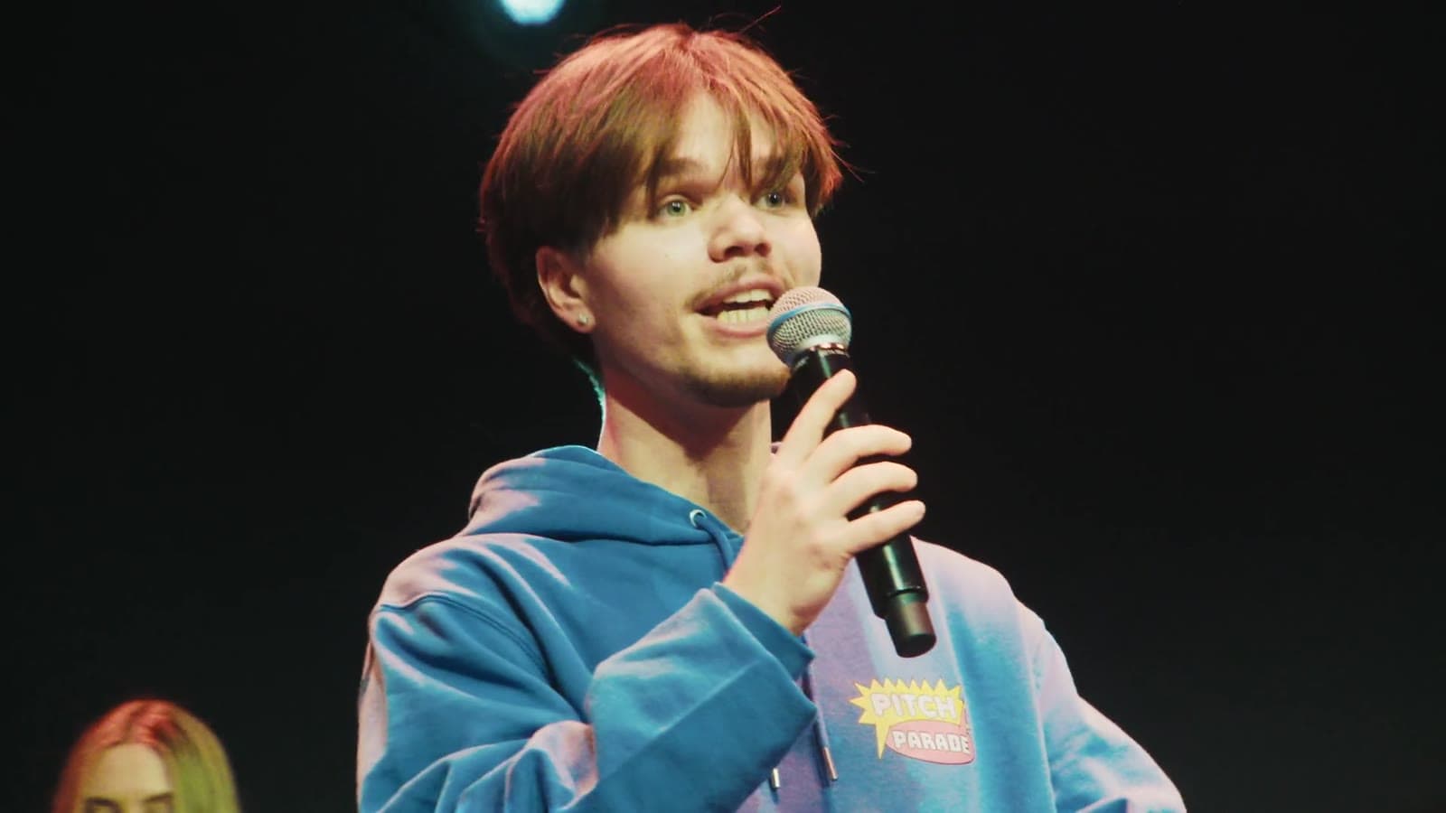 Een jongen in een blauwe hoodie pitcht met een microfoon op het podium, still uit de aftermovie van het Pitchfestival 2026
