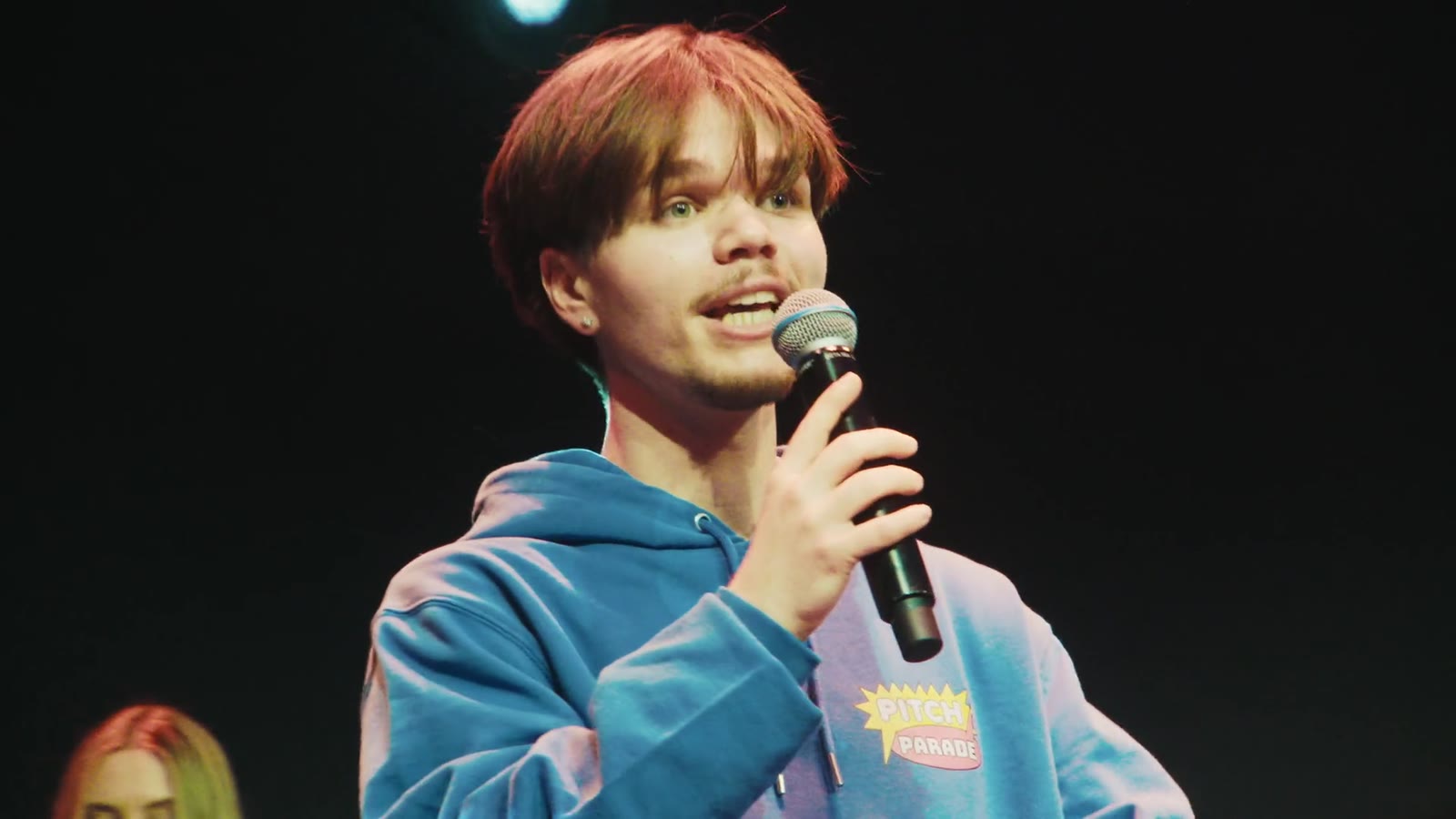 Een jongen in een blauwe hoodie pitcht met een microfoon op het podium, still uit de aftermovie van het Pitchfestival 2026
